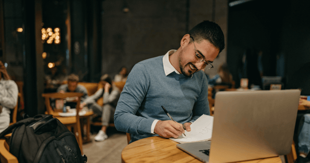 Man sitting at a table writing with a laptop in front of him