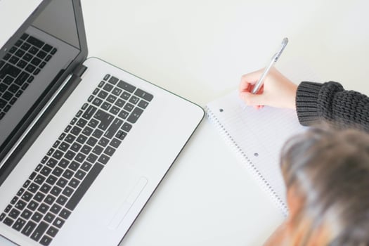 Woman taking notes with a pen and paper and looking at a laptop